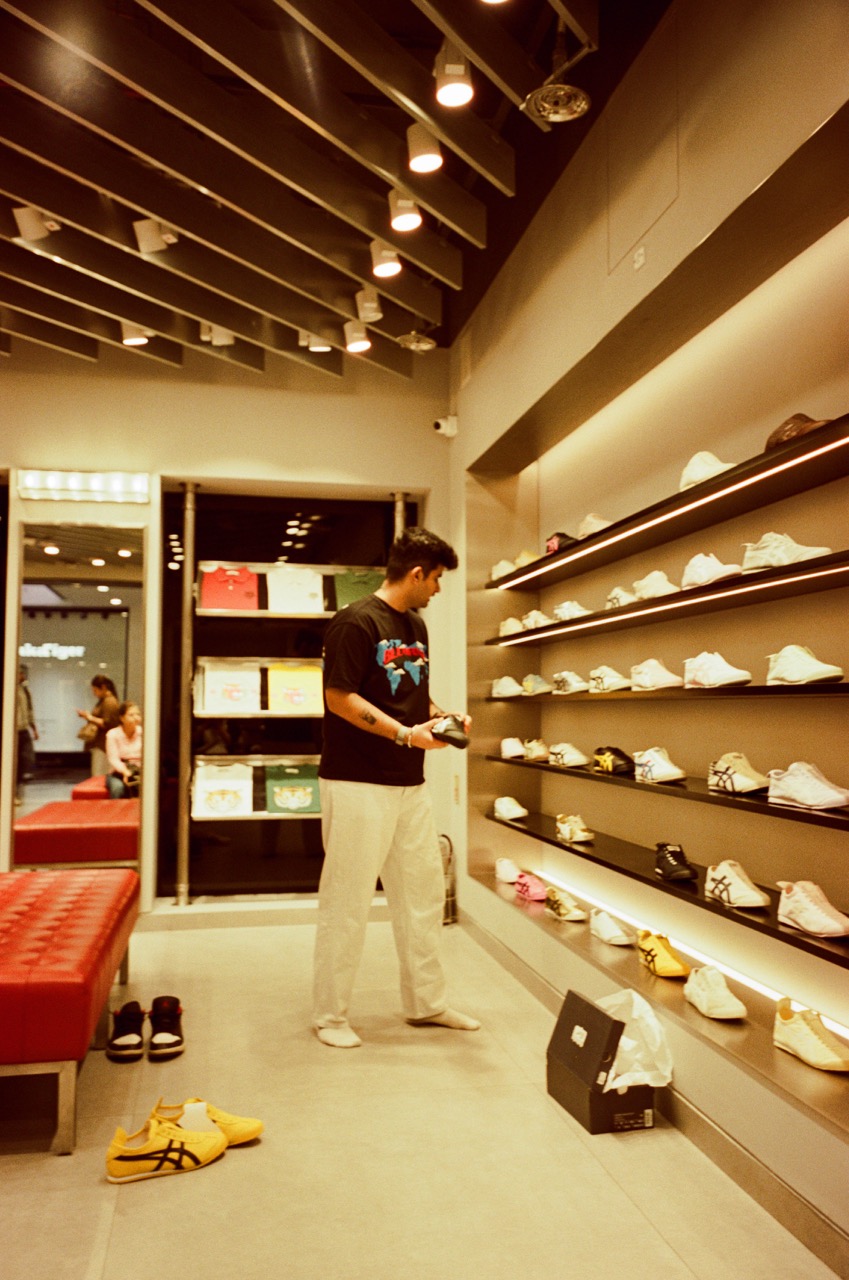 A man in a black graphic T-shirt and white trousers stands barefoot in a shoe store, looking down at a sneaker in his hand. Shelves of white and colorful sneakers line the wall beside him, lit by warm strips of light. A yellow-and-black pair sits on the floor near a red tufted bench, and a shoebox lies open on the ground.