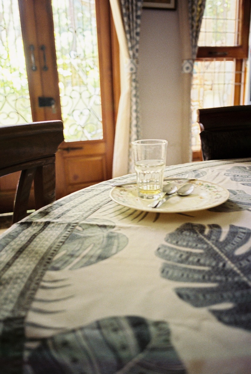 Film photo of an empty glass and a small floral plate with two spoons on a round table, with carved wooden doors and sunlit windows behind. Frontier scan: bright and airy, with clean cream tones, blue-green leaf prints on the tablecloth, and glowing, slightly blown-out windows.