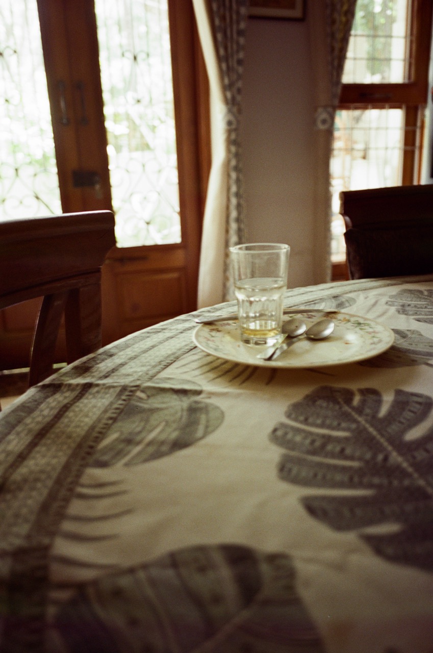 Film photo of an empty glass and a small floral plate with two spoons on a round table, with carved wooden doors and sunlit windows behind. Noritsu scan: darker and moodier, with warm olive-brown shadows, a greyer, more muted tablecloth print, and visible grain.