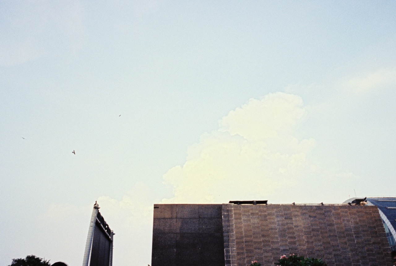 Film photo of a large cumulus cloud rising behind a stone-clad building, with a few birds in flight. Frontier scan: pale, hazy sky with a yellow-green cast in the cloud and a soft, washed-out, dreamy feel.