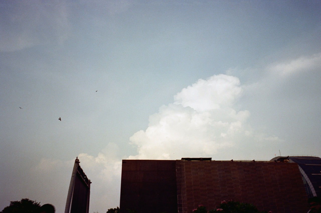 Film photo of a large cumulus cloud rising behind a stone-clad building, with a few birds in flight. Noritsu scan: deeper, cooler blue-grey sky with crisp white clouds, more contrast, and grainy, moody tones.