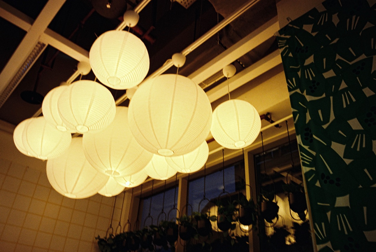 Film photo of a cluster of glowing white paper lantern lights hanging from a ceiling, with hanging plants and a green leaf-print curtain. Frontier scan: luminous, punchy yellow-gold light with vivid deep-green leaves and clean shadows.