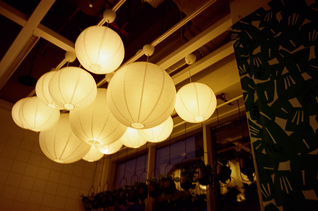Film photo of a cluster of glowing white paper lantern lights hanging from a ceiling, with hanging plants and a green leaf-print curtain. Noritsu scan: darker, amber-orange glow with heavier, muted shadows and a grainy, dusky feel.