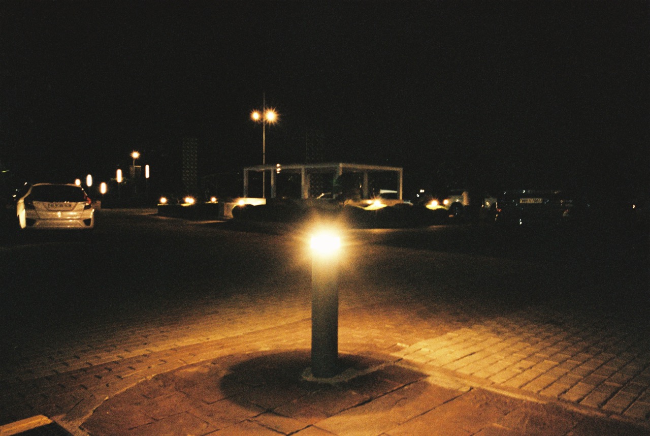 A cobblestone plaza at night, dark apart from a glowing bollard light in the foreground. A white hatchback is parked on the left, and street lamps and a pale open pavilion glow in the background.