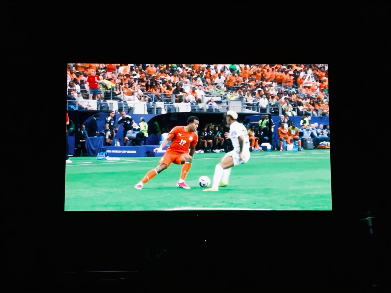 A television broadcast of a soccer match showing a player in an orange kit facing off against a defender in white on a green field, with crowded stadium stands in the background.